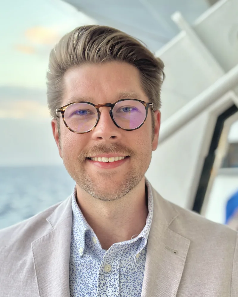 Carl Wright smiling, wearing round tortoiseshell glasses, a light linen blazer, and a blue patterned shirt, with the ocean behind him.